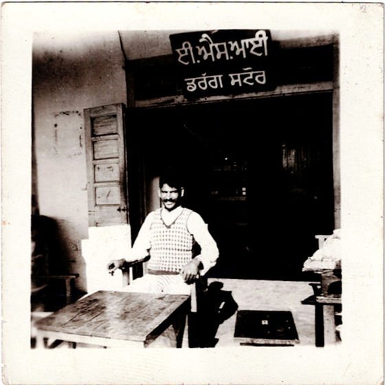 A man standing behind a wooden counter at the open front of a drug store, beneath a hand-painted Punjabi signboard reading E.S.I. Drug Store. A black-and-white print with its original border and visible age.