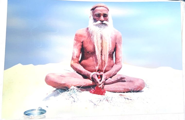 An archival photographic print, tilted slightly and worn at one corner, showing Baba Ji seated in padmasana in meditation on open pale sand beneath a wide sky.