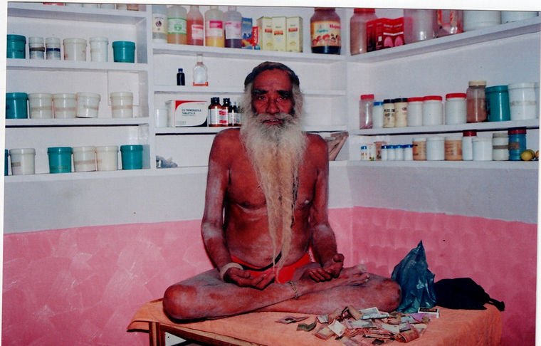 Baba Ji seated cross-legged on a wooden table inside a dispensary, surrounded on three walls by white shelves filled with jars, tubs and bottles of medicine.