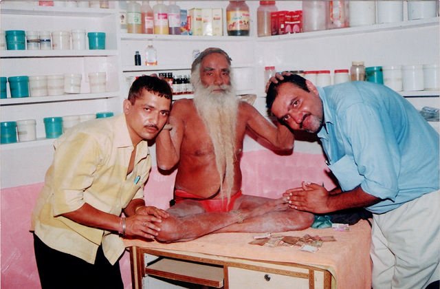 Baba Ji seated on the table in the dispensary with two men leaning in on either side of him.