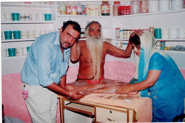 Inside the same dispensary, Baba Ji rests his hand on the head of a woman who bows towards him, while a man stands beside them.