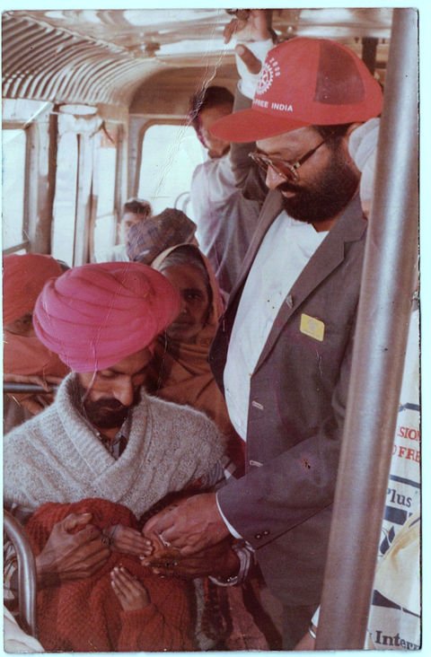 Inside a bus, a bearded man in a cap and blazer gives immunisation drops to a small child held by a man in a turban, while other passengers look on.
