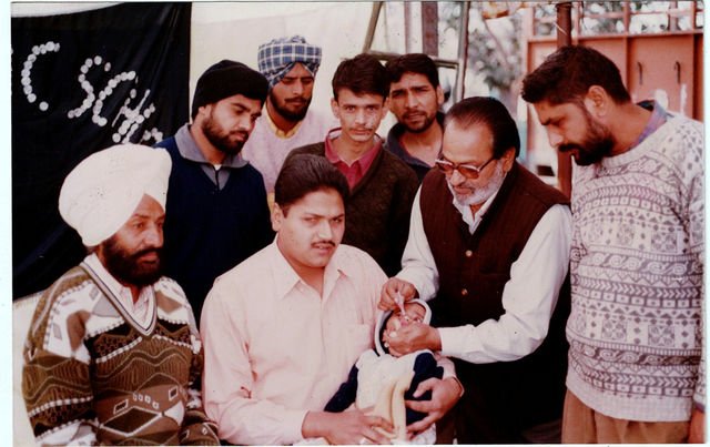 At an outdoor camp, an older man in glasses gives drops to an infant held by a seated younger man, with a group watching.