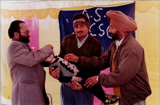 Under a decorated tent, three men together hold and attend to a child.