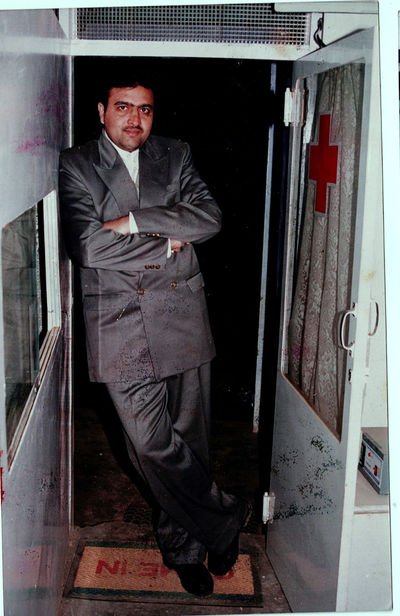A man in a grey double-breasted suit stands in a clinic doorway with his arms folded, a red cross visible on the glass door behind him.